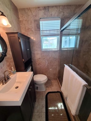 Un baño con pared de mármol, un lavamanos blanco, un inodoro, zona de ducha y una ventana con persianas.