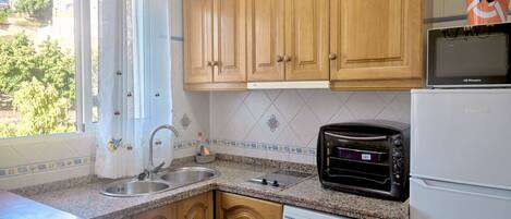 A kitchen with wooden cabinets, a microwave, a toaster, a washing machine, and a window with curtains.