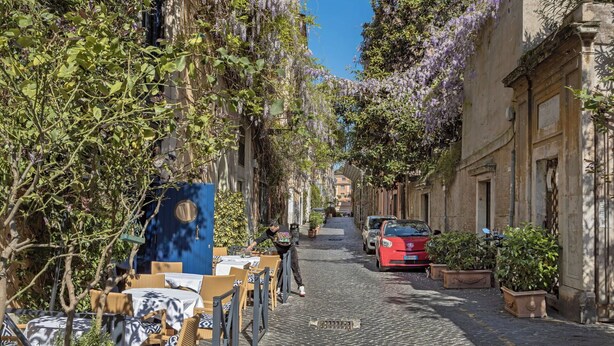 Une rue pavée bordée d’immeubles, dont certains sont recouverts de lierre et de fleurs. On y trouve des tables et des chaises en plein air, et une voiture rouge est garée sur le côté.