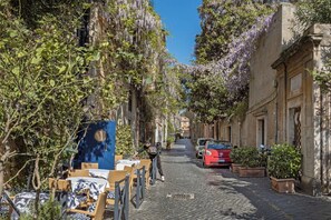 Rua de paralelepípedos ladeada por edifícios, alguns com trepadeiras e flores. Há mesas e cadeiras ao ar livre, e um carro vermelho estacionado na lateral.