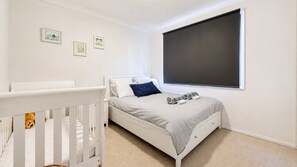 A bedroom with a crib, a bed, a window, and three framed pictures on the wall.