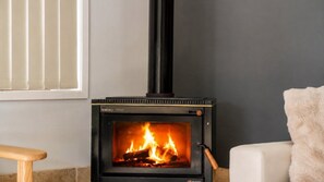 A wood-burning stove with a visible fire, a window with blinds, and a chair.