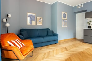 Ein Wohnzimmer mit blauem Sofa, orangem Sessel und Holzboden mit Fischgrätmuster.