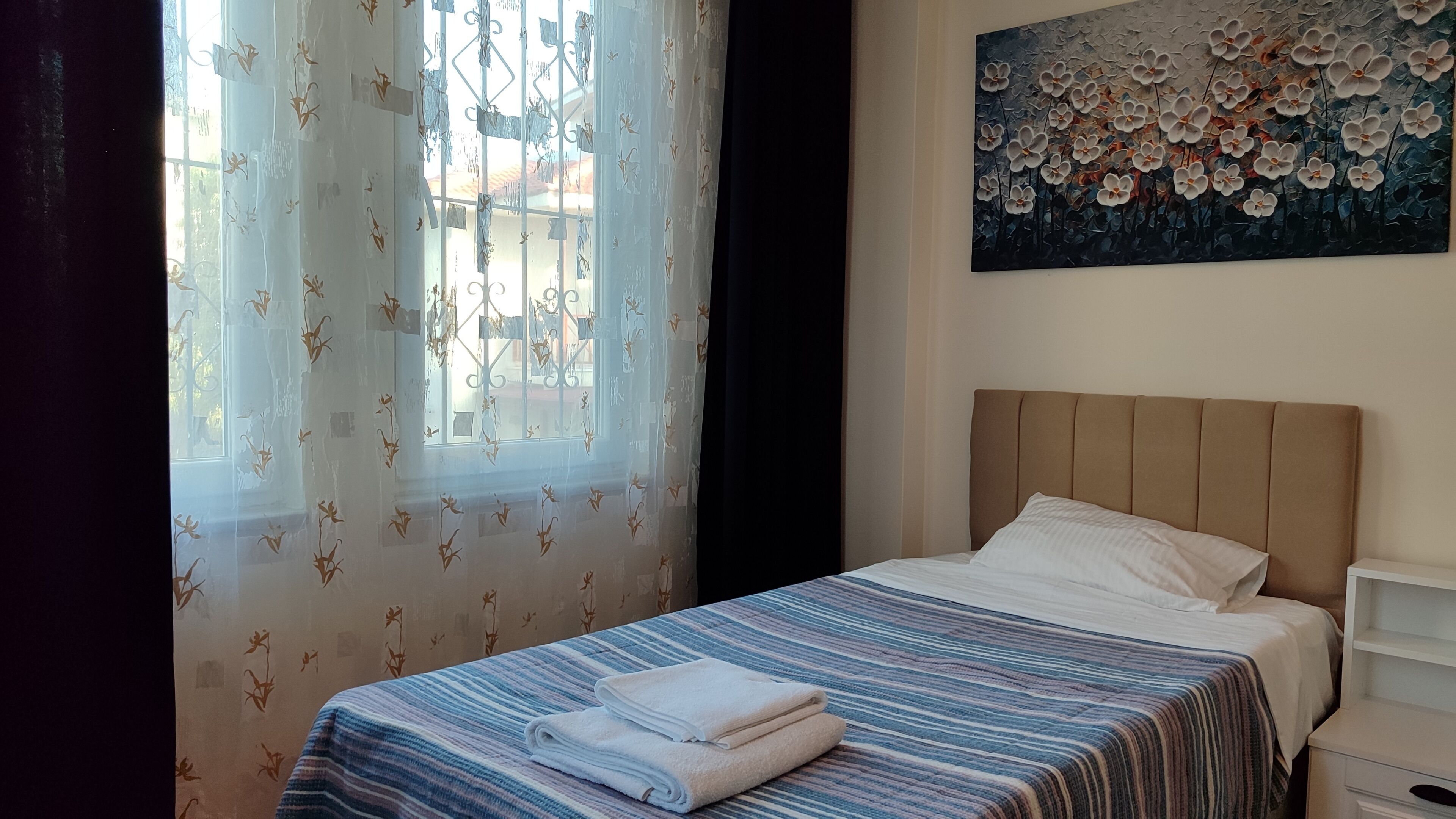 Ein Schlafzimmer mit Bett, einem Fenster mit Vorhängen, einem Bild an der Wand und einem Nachttisch.