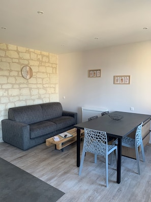 A modern living room with a grey sofa, a wooden coffee table, a dining table with chairs, and a stone wall. - 1st-floor apartment (Avoine)