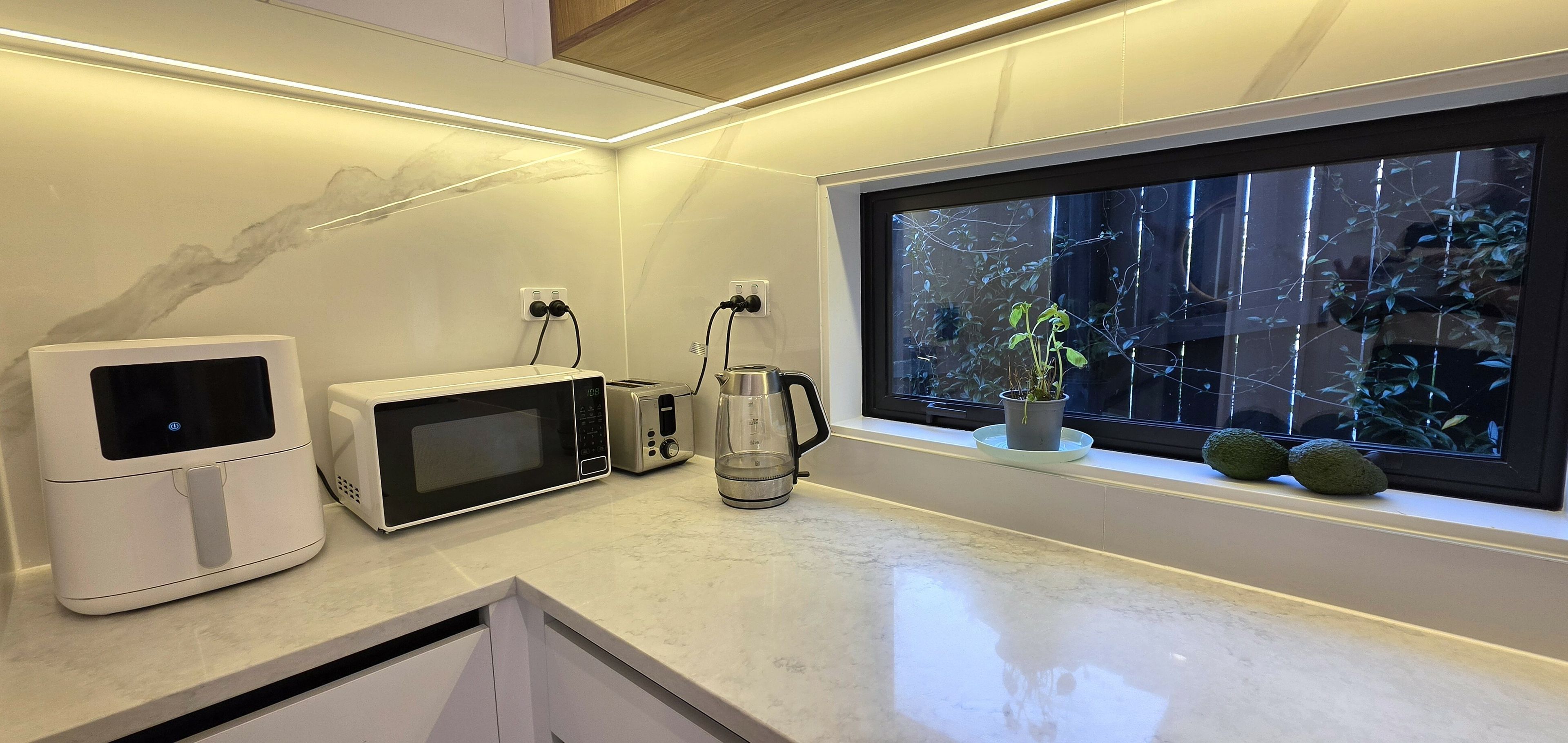 A kitchen with a microwave, toaster, and coffee maker on the counter, a window with a view of greenery, and a cabinet below.