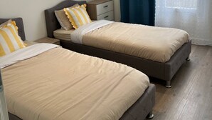 A bedroom with two beds, a nightstand, a window with curtains, and a view of buildings outside.