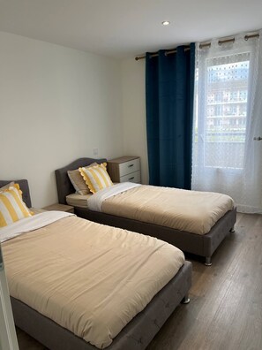 Quarto com duas camas, uma mesa de cabeceira, uma janela com cortinas e vista para edifícios.