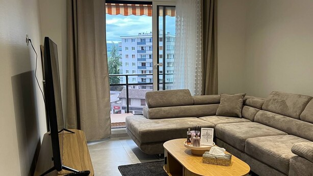 A modern living room with a sofa, a coffee table, a TV on a stand, and a view of a building through large windows.