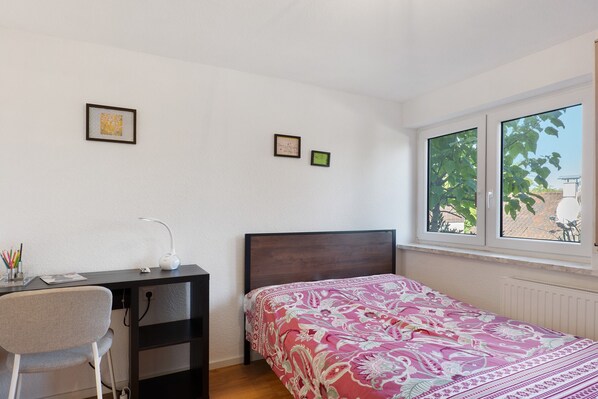 A bedroom with a bed, a desk, and a chair. There are two framed pictures on the wall.