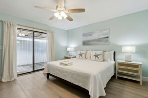 A bedroom with a bed, two bedside tables, a ceiling fan, a view through a sliding glass door, and a painting on the wall.