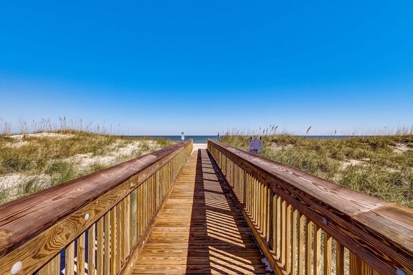 Une promenade en bois qui mène à une plage bordée de dunes de sable, sous un ciel bleu et dégagé.