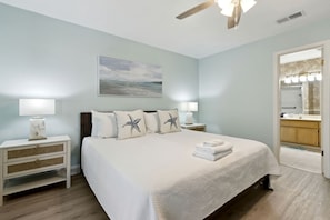 A bedroom with a bed, nightstands, a ceiling fan, a painting, and a bathroom visible through an open door.