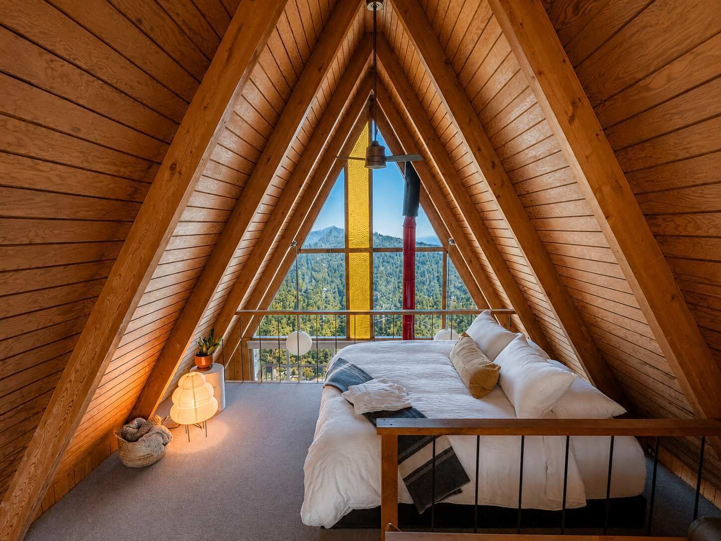 Ein Zimmer mit hohen, giebelförmigen Holzwänden, einem Bett, einem kleinen Tisch und Blick auf eine bewaldete Bergkette.