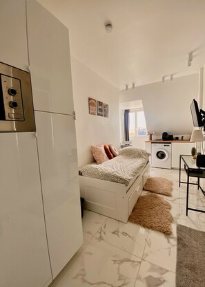 A compact, modern room with a bed, a washing machine, a desk with a computer, and a window with curtains. - Cozy Studio in Neuilly, Bois de Boulogne, and La Défense (Neuilly-sur-Seine)