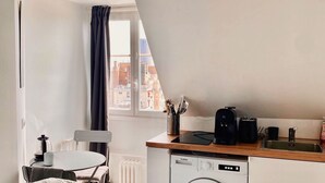 Een knus woongedeelte met een bed, een eethoek, een keuken met wasmachine, en uitzicht op de stad door een raam.