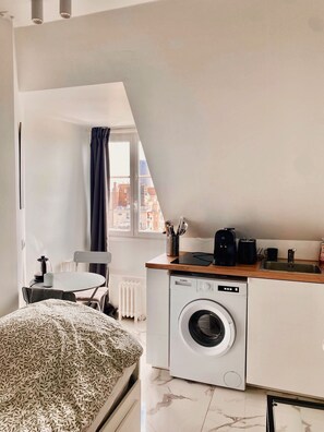 A compact living space with a bed, a dining area, a kitchen with a washing machine, and a view of the city through a window. - Cozy Studio in Neuilly, Bois de Boulogne, and La Défense (Neuilly-sur-Seine)