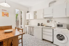 Una cucina compatta con mobili bianchi, un forno a microonde, una lavatrice e un'area pranzo con un tavolo e sedie in legno.