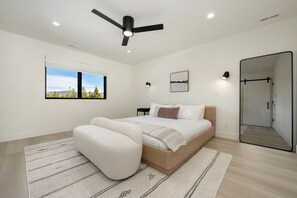 Un dormitorio moderno con una cama, una silla, un ventilador de techo, una ventana con vistas, una mesita de noche y un espejo.