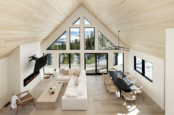 A modern interior with a high ceiling, large windows, a fireplace, and a dining area.