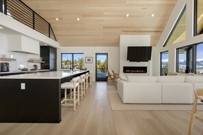 A modern kitchen with a central island, a dining area with chairs, a living room with a sofa, a fireplace, and large windows with a mountain view.