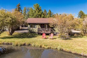 A cabin with a deck, two Adirondack chairs, a picnic table, and a pond in front.