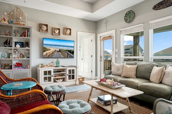 A living room with a large TV, wicker furniture, a bookshelf, and a view of the ocean.