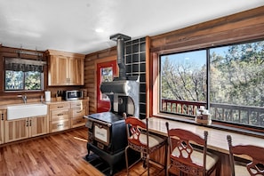 A cabin interior with a wood stove, wooden cabinets, a sink, and a dining area with a view of trees.