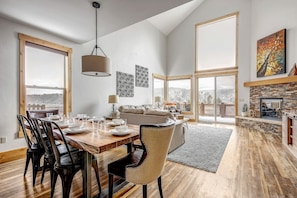 A spacious living area with a dining table, chairs, a sofa, a fireplace, and large windows offering a view.
