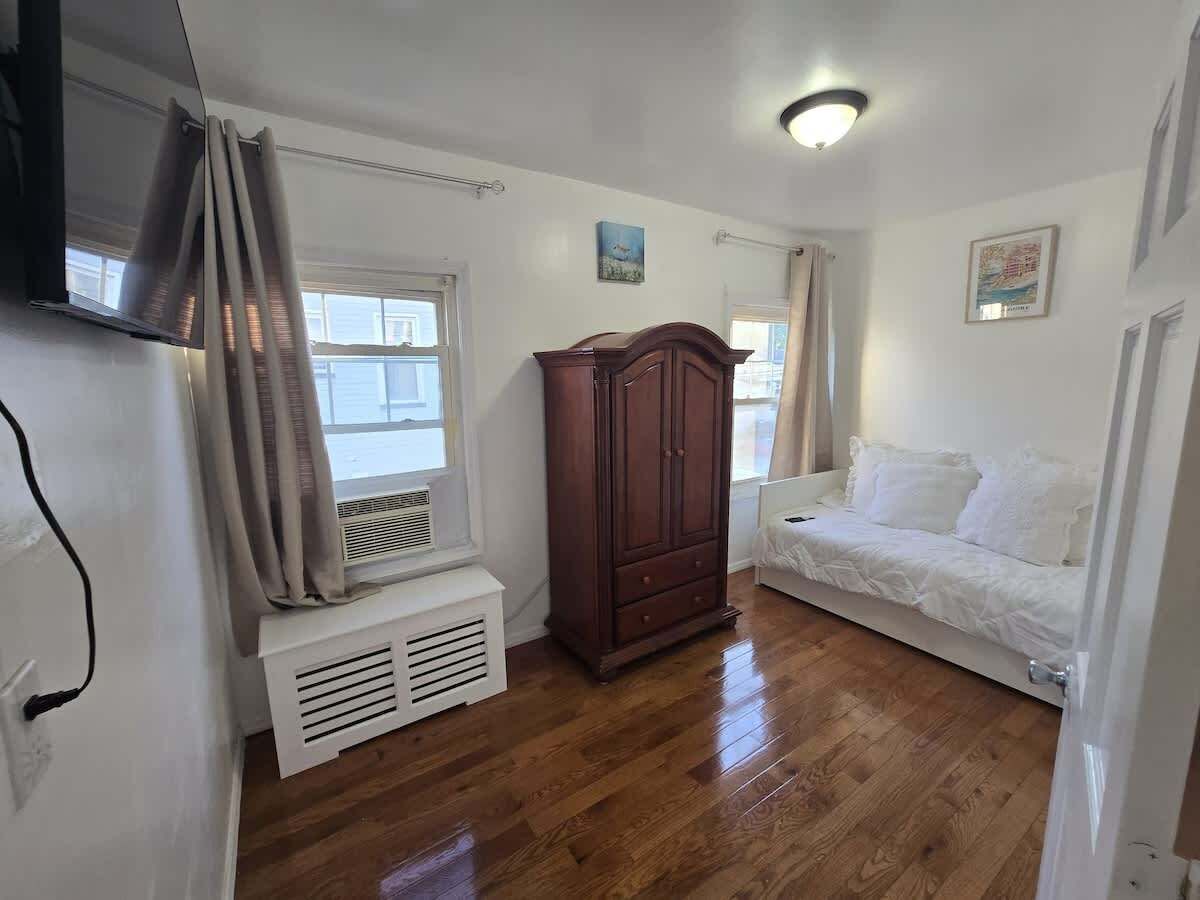 Una habitación con piso de madera, un radiador blanco, un armario de madera, un sofá blanco y una ventana con cortinas.