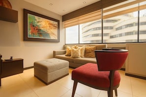 Ein modernes Wohnzimmer mit einer Couch, einem Hocker und einem roten Sessel, einem Gemälde an der Wand und Blick auf ein Gebäude durch große Fenster.