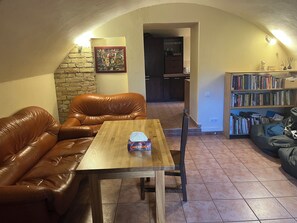 A cozy living room with a leather couch, a wooden table, a bookshelf, and a kitchen in the background.