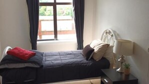 Um quarto com cama, mesa de cabeceira com luminária, janela com cortinas e uma pequena planta.