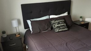 Um quarto com cabeceira escura, cama com travesseiro marrom e branco, mesa de cabeceira com abajur e um letreiro na parede listando cidades.