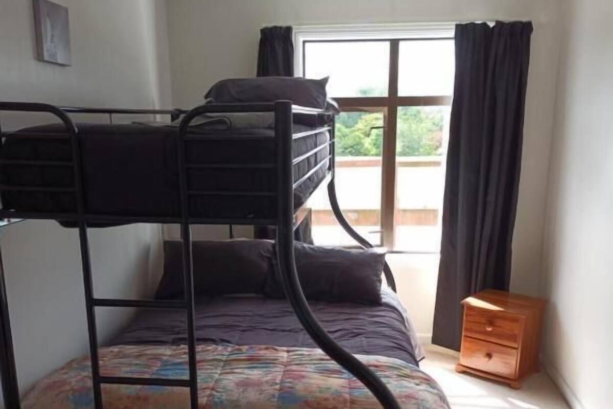 Ein Etagenbett mit einer bunten Decke, ein Fenster mit Vorhängen und ein kleiner Nachttisch aus Holz.