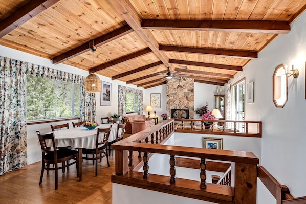 A dining area with wooden beams, a stone fireplace, and a view of greenery outside.