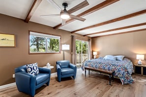 A bedroom with a bed, two chairs, a table, a ceiling fan, and a window with a view of trees.