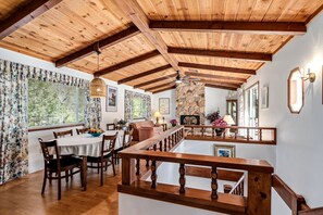 A dining area with wooden beams, a stone fireplace, and a view of greenery outside.