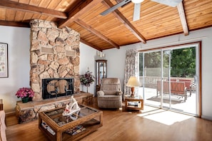 A living room with a stone fireplace, wooden ceiling beams, a glass door leading to an outdoor patio, and a comfortable seating area.
