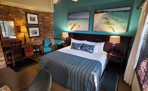 Habitación de hotel con pared de ladrillo, cama con ropa de cama estampada, escritorio con lámpara y dos cuadros en la pared.