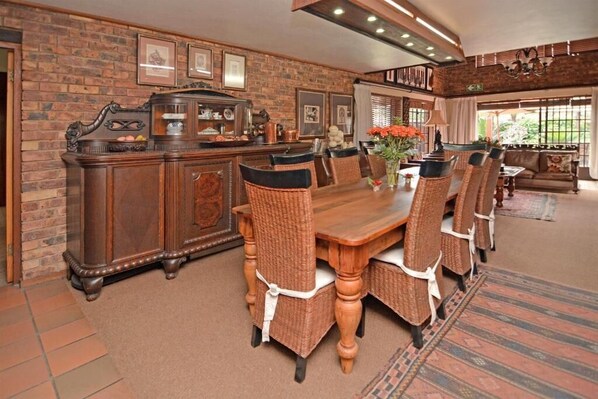A dining area with a wooden table and wicker chairs, a brick wall with framed pictures, and a large window with a view.