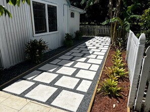 Un chemin mène à un bâtiment blanc doté d’une fenêtre et d’une porte, entouré d’une clôture en piquets blancs et de diverses plantes.