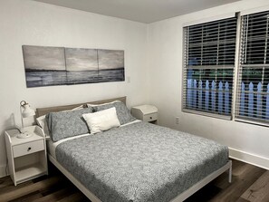 Un dormitorio con cama, mesitas de noche, un cuadro colgado en la pared y una ventana con persianas.
