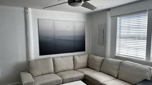 A living room with a beige sectional sofa, a white coffee table, a ceiling fan, a large abstract painting, and a window with blinds.