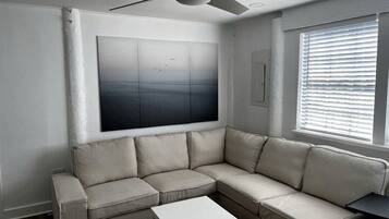 Un salon avec un canapé d’angle beige, une table basse blanche, un ventilateur de plafond, un grand tableau abstrait et une fenêtre avec des stores.