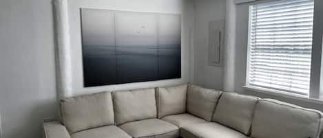 A living room with a beige sectional sofa, a white coffee table, a ceiling fan, a large abstract painting, and a window with blinds.
