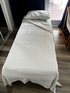 A single bed with a white bedspread in a room with wooden flooring and a window.