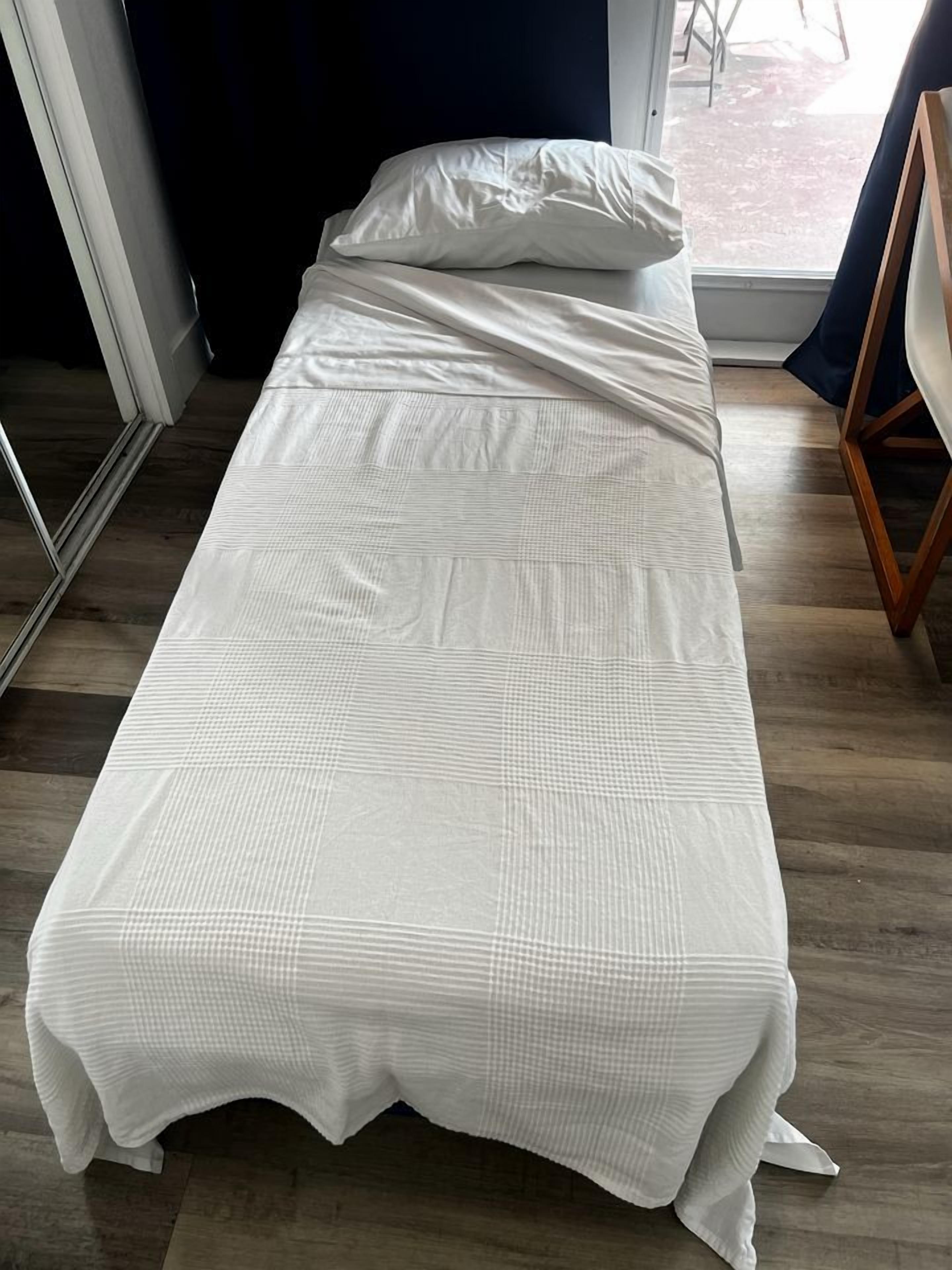 A single bed with a white bedspread in a room with wooden flooring and a window.