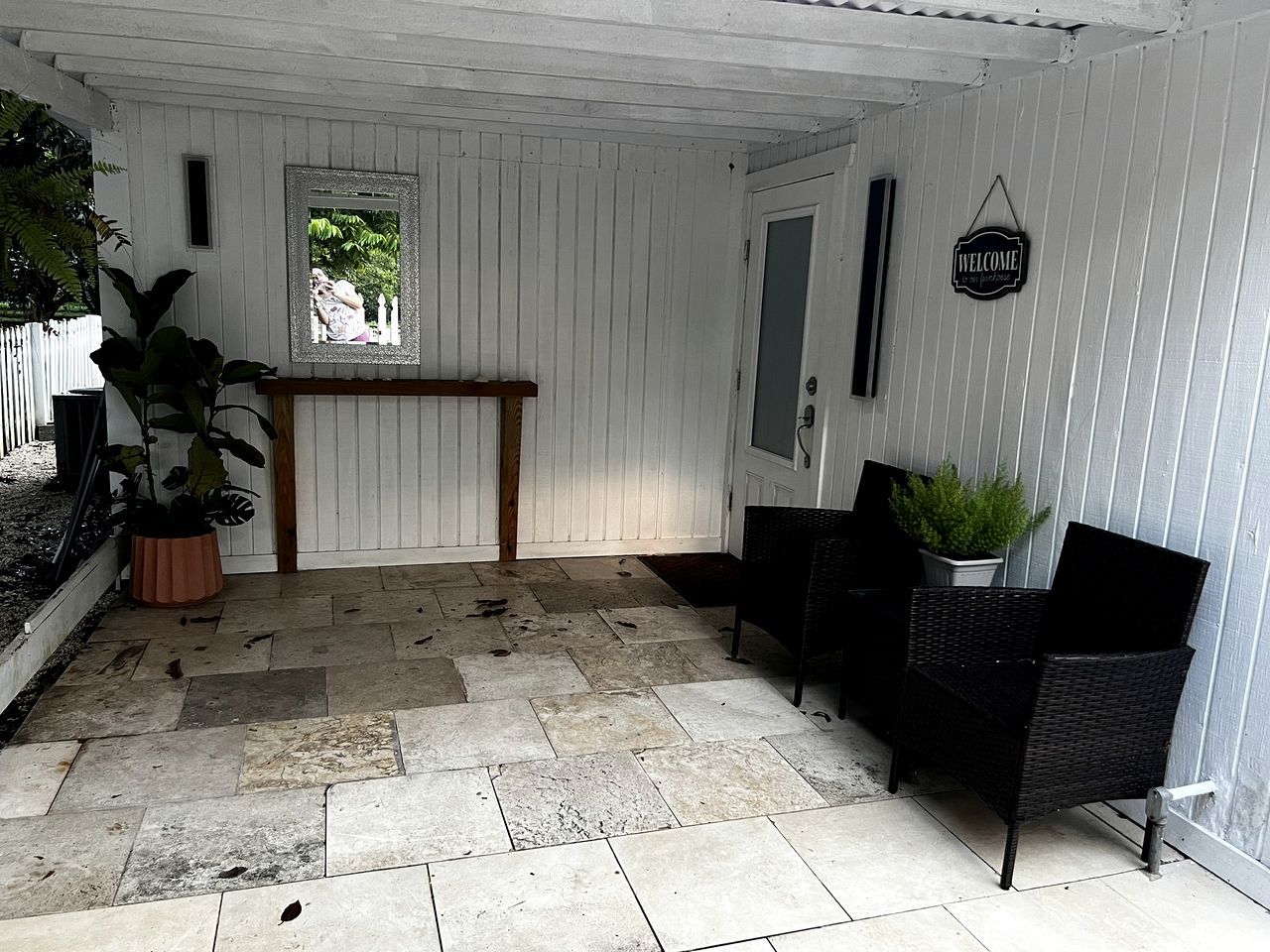 Un patio cubierto con suelo de piedra, un banco de madera, sillas de mimbre negro y un cartel de bienvenida.