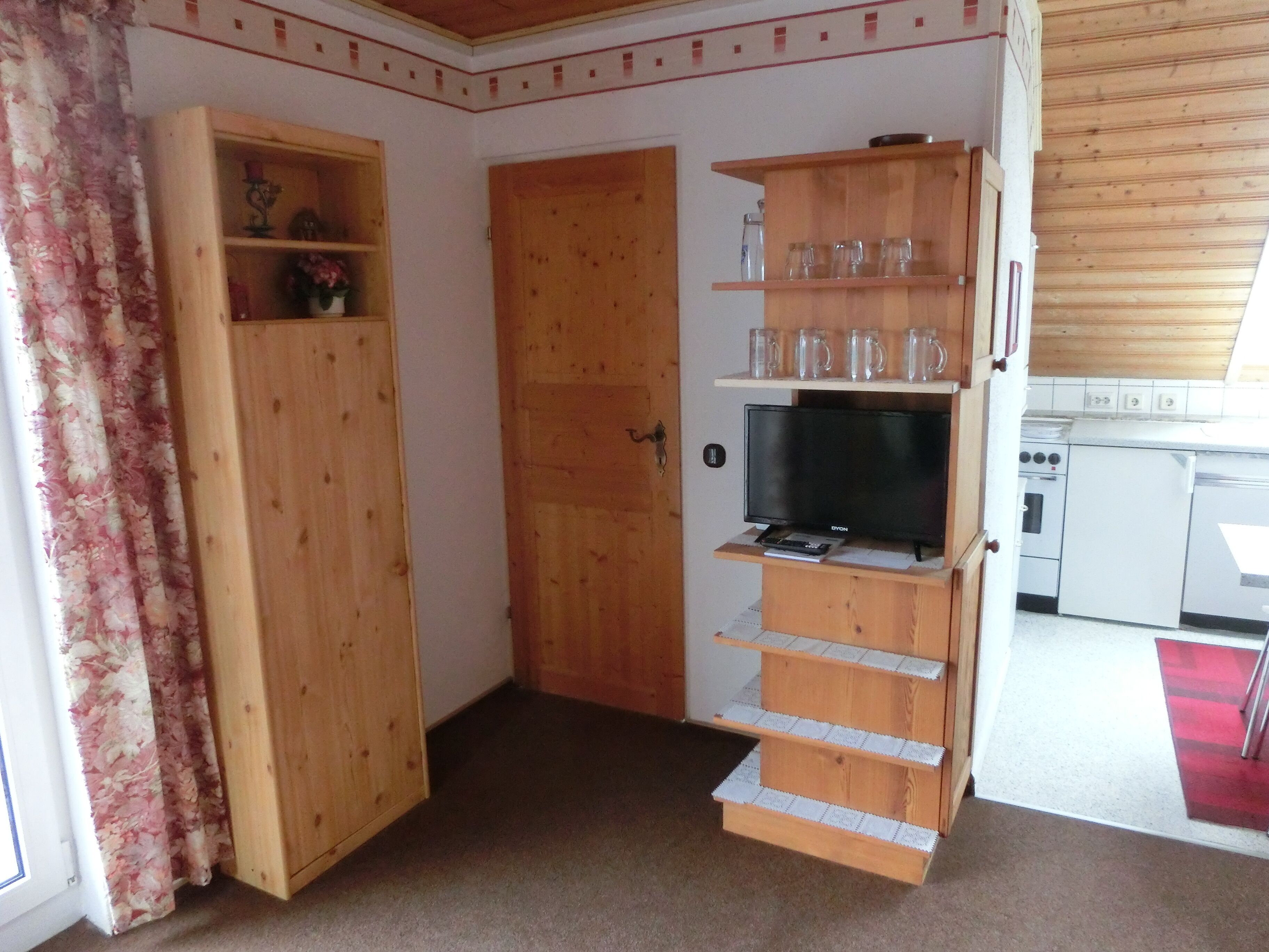 Ein kompakter Wohnraum mit Holztür, Fernseher auf einem Standfuß und Küchenzeile mit weißen Geräten.